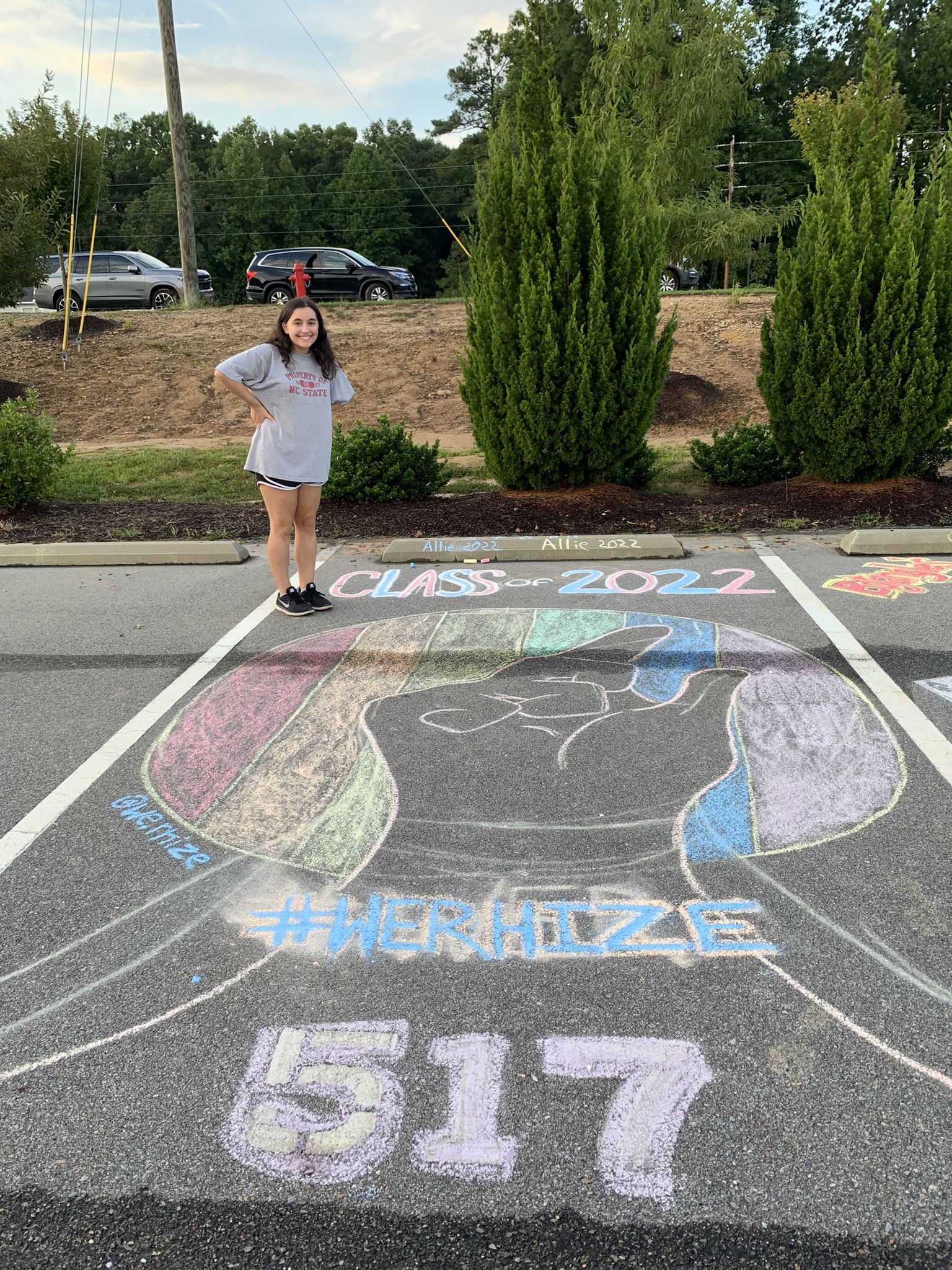 Girl standing next to chalk art. The chalk art is a picture of a hand infront of a rainbow flag. Below the flag it says says #WERHIZE 517.